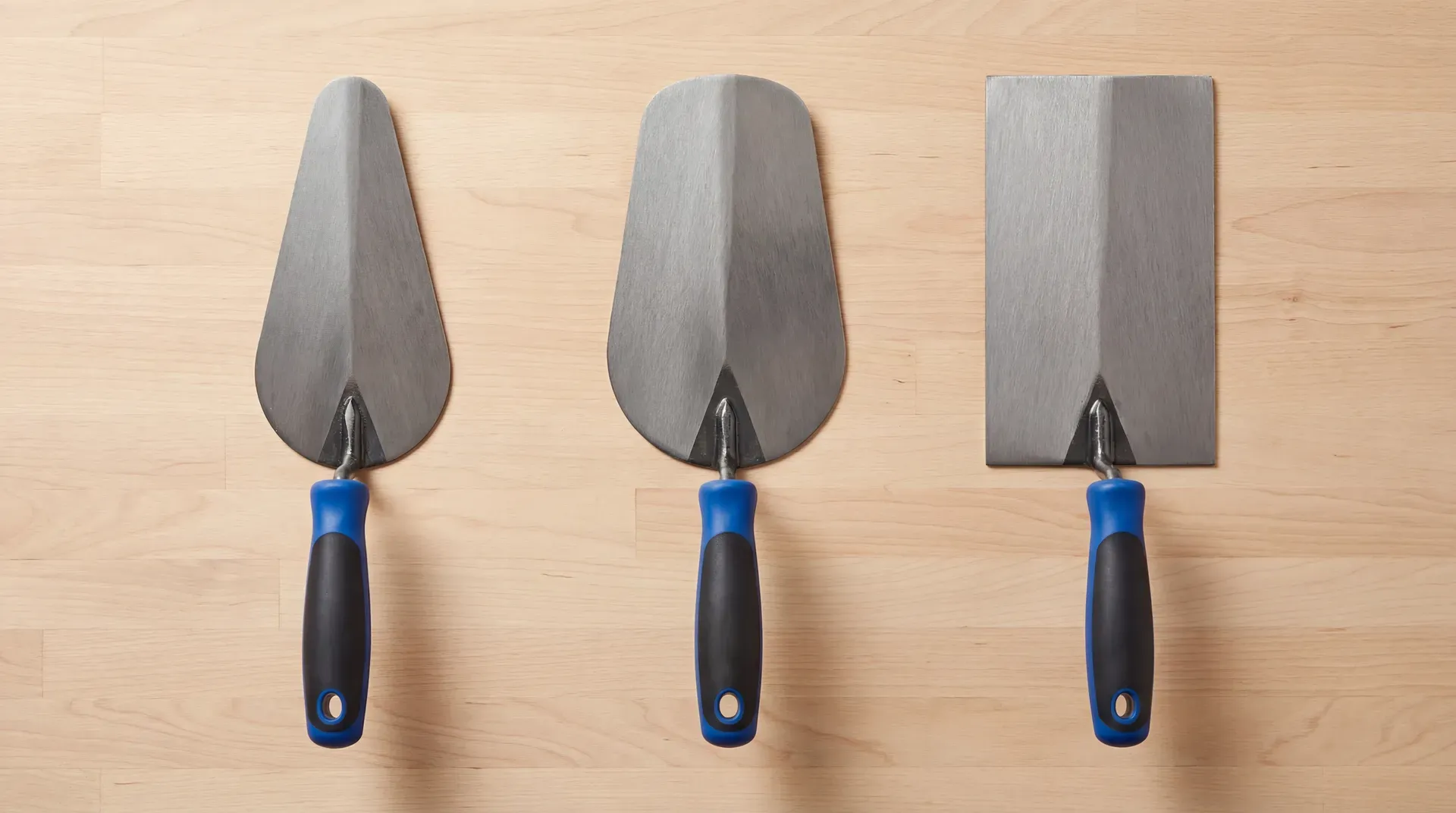 Three brick trowels laid side by side showing the London pattern with a rounded heel and narrow blade on the left, Wide London with a broader heel in the centre, and Philadelphia with a square heel and widest blade on the right.