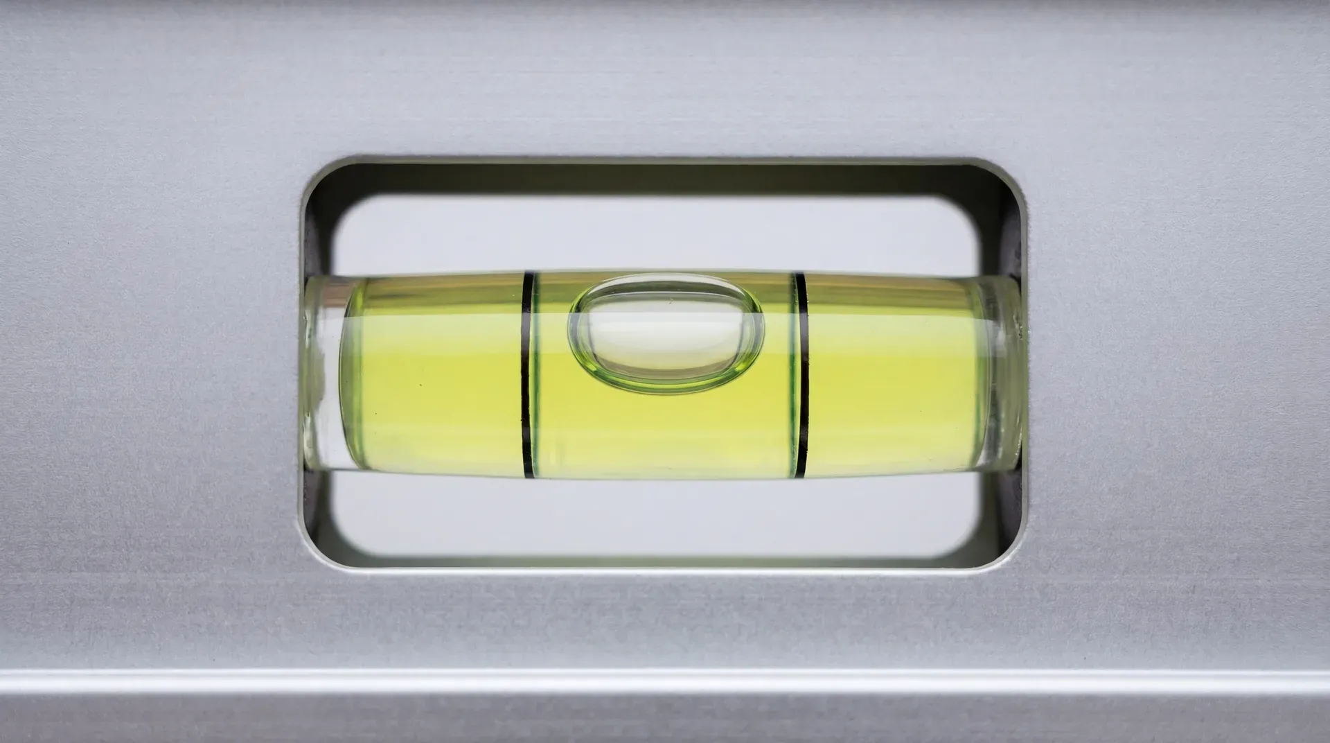 Extreme close-up of a spirit level bubble vial photographed from directly above, showing the yellow-green liquid, the air bubble centred between the two etched reference lines, set into a silver-grey aluminium body with rectangular vial window.