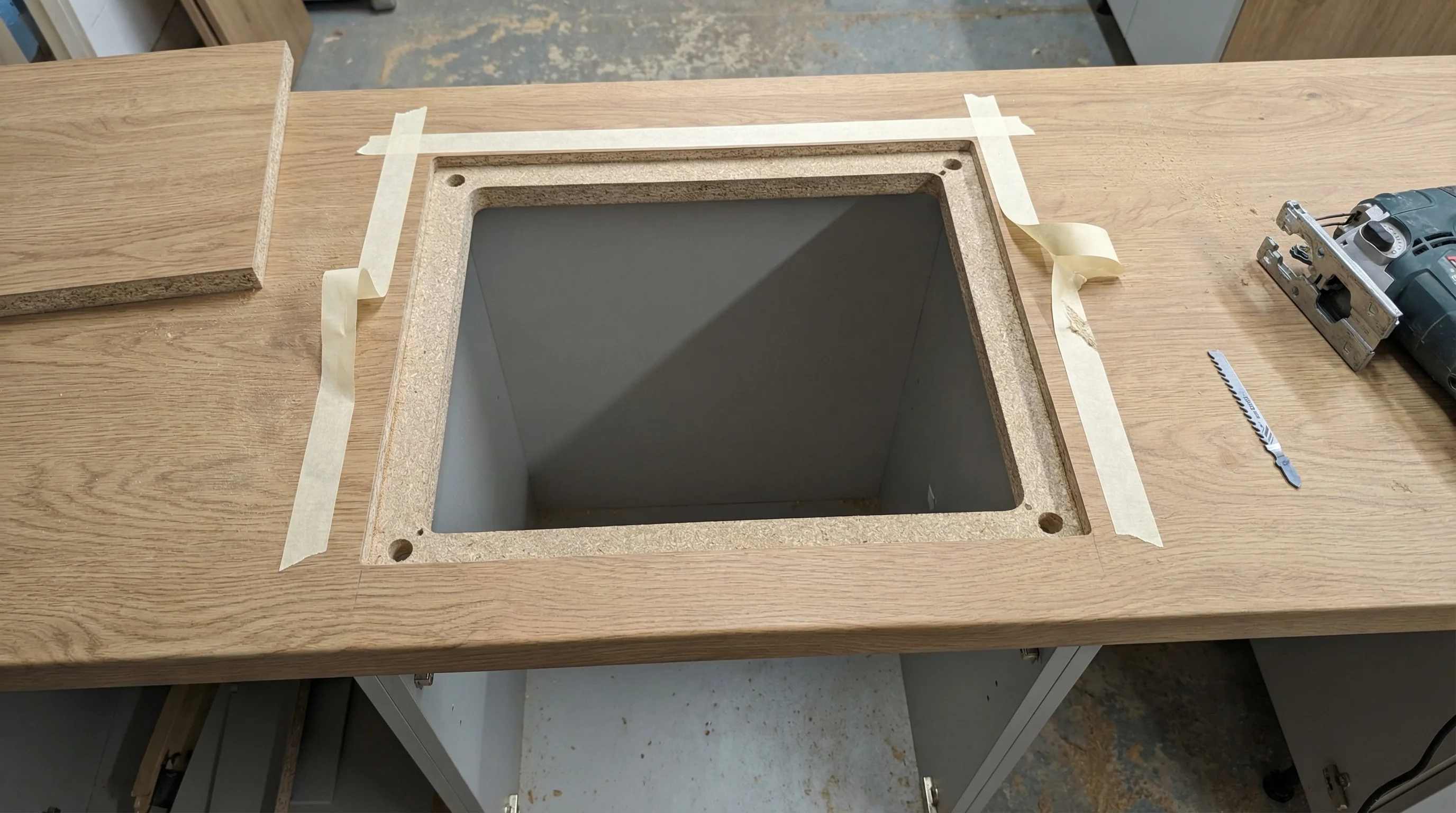 Overhead view of a laminate worktop with a cleanly cut rectangular sink cutout. The cut edges show no chipping or tear-out. Masking tape is partially peeled back, the waste piece sits beside the worktop, and a caulking gun applies sealant to a cut edge. Drilled pilot holes are visible at each corner.